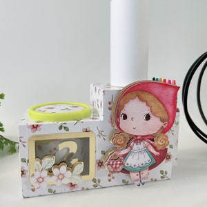 Puede incluir: Una caja de madera blanca con un patrón floral y un personaje de niña con capucha roja. La caja tiene una ventana transparente con el número "2" en su interior. Hay un cilindro blanco en la parte superior de la caja con una fila de lápices de colores.