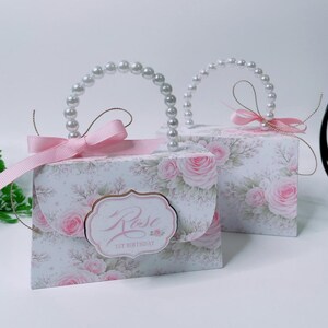Puede incluir: Dos cajas de regalo rectangulares con forma de bolso, fondo blanco y estampado de rosas rosas. Cada caja tiene un asa de perlas blancas, un lazo de cinta rosa y una etiqueta con el texto Rose 1ST BIRTHDAY.