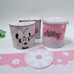 May include: Two pink and white cylindrical containers with Minnie Mouse designs. One container has a white lid and the other has a pink lid. The containers have white polka dots and white flowers. The container with the pink lid has the name "Ashley" written on it. A pink and white patterned paper with white flowers is in the background.