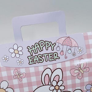May include: A pastel pink and white Easter-themed gift bag with a handle. The bag features a gingham pattern and illustrations of flowers, butterflies, and a bunny. The words "HAPPY EASTER" are printed on the bag.