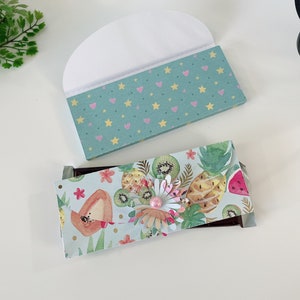May include: Two rectangular chocolate bar wrappers. One is teal with yellow stars and pink hearts. The other features a tropical fruit pattern with pineapples, kiwi, and watermelon, accented by a layered paper flower with a central pink bead.