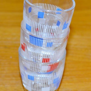 May include: A stack of four clear glass shot glasses. Each glass is decorated with a white vertical striped pattern and accented with red, blue, and white squares. The glasses are stacked on top of each other.