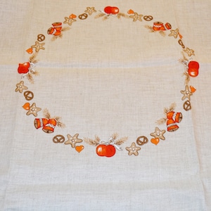 May include: A beige tablecloth with a brown and orange wreath design. The wreath is made up of gingerbread cookies, apples, and bells.