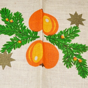 Op de afbeelding: Een witte jute stof met een groene dennentak en drie oranje ornamenten. De ornamenten zijn in de vorm van een ster en twee cirkels. Er zijn twee bruine sterren in de hoeken van de stof.
