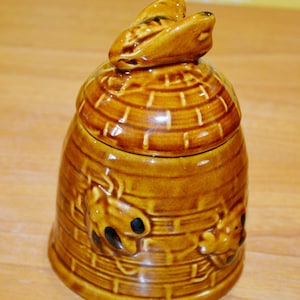 Könnte beinhalten: Ein brauner Keramik-Honigtopf in Form eines Bienenstocks mit einer Biene oben drauf. Der Topf hat eine strukturierte Oberfläche und ist mit Bienen verziert.