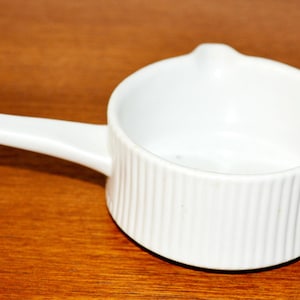 May include: A white ceramic sauce warmer with a handle and a pouring spout. The small, round bowl has vertical ribbed detailing. The handle extends from the side of the bowl. The warmer sits on a wooden surface.