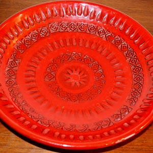 May include: A circular, red ceramic plate with a raised, textured design. The plate features concentric circles of geometric patterns and a central floral motif. The rim has teardrop-shaped indentations. The plate is a vibrant shade of red.