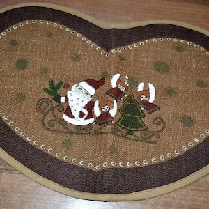 May include: A brown burlap Christmas tree skirt with a dark brown border. The skirt features a hand-painted design of Santa Claus and three angels pulling a sleigh with a Christmas tree.