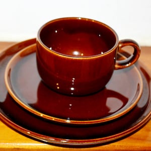 Puede incluir: Un juego de vajilla de cerámica de tres piezas en un color marrón oscuro intenso. El juego incluye una taza con asa y dos platos. La taza y los platos tienen un acabado brillante y están colocados sobre una superficie de madera.