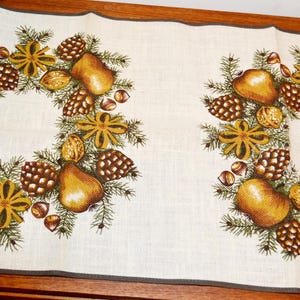 May include: A white linen table runner with two embroidered wreaths featuring pears, pine cones, nuts, and star anise. The wreaths are in shades of brown, green, and gold.