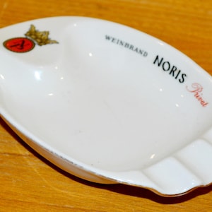 Op de afbeelding: Witte keramische asbak met een tweedelig ontwerp. De asbak heeft de woorden "WEINBRAND NORIS Privat" erop gedrukt, samen met een rood en goudkleurig embleem. De asbak heeft een gouden rand.