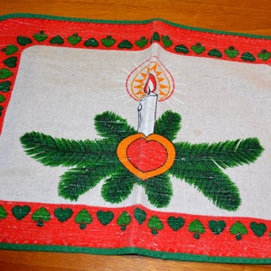 May include: A red and green Christmas table runner with a white background featuring a candle and a heart in a wreath of green pine branches.