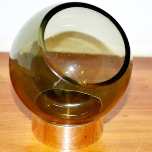 Può includere: Una ciotola sferica in vetro verde oliva con una grande apertura e una base in metallo color argento. La ciotola ha un'apertura circolare sul lato. Il vetro ha una leggera tinta e la base ha una forma arrotondata.