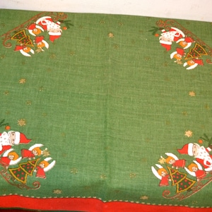 May include: A green Christmas table runner with a red border. The runner features embroidered images of Santa Claus in a sleigh with angels and Christmas trees. Gold star accents are scattered across the green fabric.