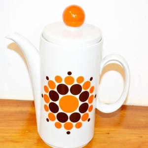 Puede incluir: Cafetera de cerámica blanca con tapa y asa. La cafetera presenta un diseño de círculos naranjas y marrones. La tapa está rematada con una pequeña esfera naranja. La cafetera tiene un pico largo.