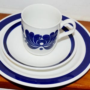 Op de afbeelding: Een witte keramische mok en bordenset met een blauw bloemmotief. De mok heeft een oor en staat op een klein bord, dat op een groter bord staat. De borden hebben een blauwe rand.