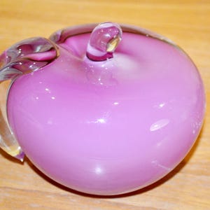 May include: A decorative glass apple in shades of pink and purple. The apple has a stem and a curved leaf-like handle. The glass is smooth and glossy, reflecting light. The apple is sitting on a wooden surface.