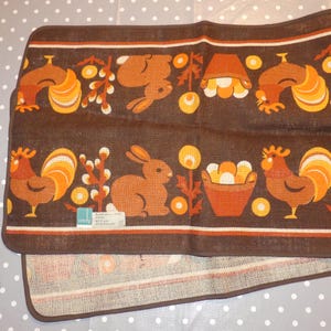 Può includere: Asciugamano da cucina marrone e arancione con un motivo ripetitivo di galli, conigli e cesti di uova. L'asciugamano ha un bordo marrone e il testo "andy" in bianco.