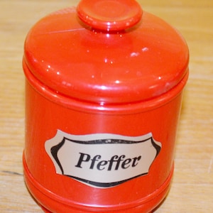 Peut inclure: Un pot à épices rouge vif avec un couvercle assorti et une étiquette indiquant "Pfeffer". Le récipient est cylindrique avec un dessus et une base légèrement arrondis. L'étiquette est blanche avec un bord noir.