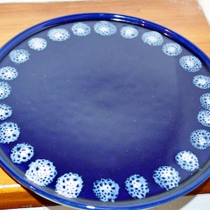 May include: A round, deep blue ceramic plate with a dark blue rim. The plate features a ring of white, floral-like patterns around the edge. The plate is on a wooden surface.