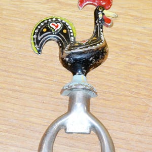 May include: A decorative bottle opener featuring a black rooster with red, yellow, and green accents. The rooster has a heart design on its wing and sits atop a silver bottle opener.