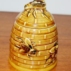 Può includere: Un vasetto di miele in ceramica a forma di alveare, con coperchio e una figura di ape sulla parte superiore. Il vasetto è marrone con una trama intrecciata e presenta decorazioni a forma di ape. È un oggetto decorativo per la cucina.