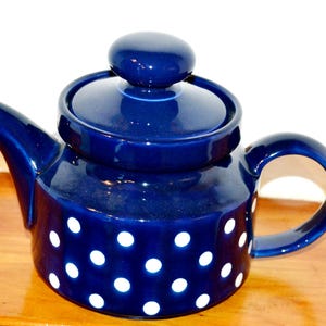 May include: A dark blue ceramic teapot with a lid and handle. The teapot is adorned with white polka dots. The spout and handle are curved. The teapot is on a wooden surface.