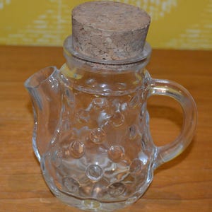 Peut inclure: Une carafe en verre transparent avec un bouchon en liège. La carafe a une poignée et un bec verseur, et est ornée de petits cercles en relief. Le bouchon en liège est de couleur marron clair. La carafe est posée sur une surface en bois.