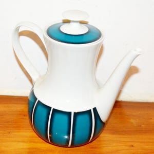 Puede incluir: Una tetera de cerámica blanca con un diseño turquesa y blanco. La tetera tiene un asa curva, un pico y una tapa con un pequeño pomo. La parte inferior de la tetera presenta rayas verticales turquesas con detalles blancos.