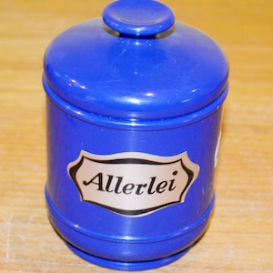 Peut inclure: Un récipient bleu cobalt avec un couvercle et une étiquette décorative. L'étiquette porte le mot "Allerlei" dans une police stylisée. Le récipient est cylindrique avec un dessus arrondi et un petit bouton sur le couvercle.