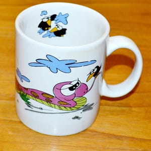 Puede incluir: Taza de cerámica blanca con una ilustración de dibujos animados de una serpiente rosa con ojos negros y un pájaro negro con un pico largo. La serpiente está en una hoja verde y el pájaro está volando en el cielo. Hay tres pájaros negros en el cielo.