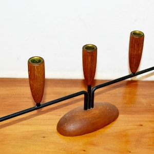 May include: A mid-century modern candle holder with five wooden candle cups and a black metal arm. The base is oval-shaped and made of wood. The candle cups have brass-coloured interiors. This decorative piece is designed to hold candles.