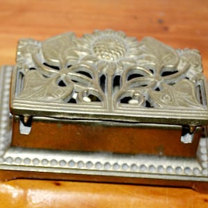 May include: A vintage brass match safe featuring a sunflower and leaf design on the lid. The rectangular box has a decorative beaded border and a hinged lid. The match safe is made of brass and sits on a wooden surface.