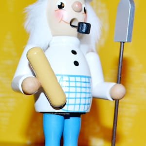 Könnte beinhalten: Eine weiße Holzfigur eines Bäckers, der einen weißen Hut trägt und ein Brotlaib und einen Holzspatel hält. Der Bäcker trägt eine blau-weiß karierte Schürze und eine blaue Hose.