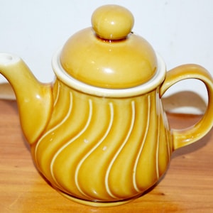 Könnte beinhalten: Eine gelbe Keramik-Teekanne mit Deckel und gebogenem Griff. Die Teekanne hat einen weißen Rand und vertikale, wellenförmige Linien. Der Ausguss ist weiß. Die Teekanne steht auf einer Holzoberfläche.