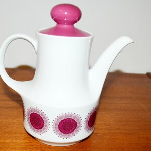 Könnte beinhalten: Weiße Keramik-Kaffeekanne mit magenta Deckel und Griff. Die Kanne hat ein dekoratives Muster aus magentafarbenen Kreisen am Boden. Der Ausguss ist gebogen und der Deckel hat einen runden Knopf. Ein Vintage-Küchenartikel.