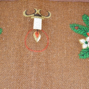 May include: A brown burlap fabric with a red and green poinsettia design. The fabric has a light brown background with a woven texture. The poinsettia flowers are red with green leaves and white centers. There is a gold candle holder with a white candle in the center of the fabric.