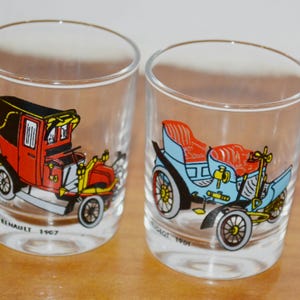 Könnte beinhalten: Zwei transparente Gläser mit Illustrationen von Oldtimern. Eines zeigt einen roten und schwarzen Renault von 1907, das andere einen blauen und roten Peugeot von 1901. Beide Gläser haben goldene Ränder und stehen auf einer Holzoberfläche.