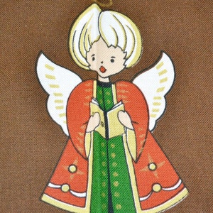 May include: A fabric Christmas angel decoration with a halo, white hair, and wings. The angel wears a red robe with gold trim over a green dress and holds a book. The background is a solid brown color.