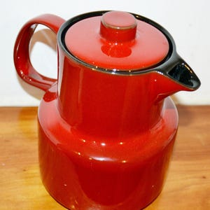 Puede incluir: Una cafetera de cerámica roja con borde y asa negros. La cafetera tiene una tapa con un pequeño pomo y un pico para verter. La cafetera probablemente se utiliza para preparar y servir café o té.