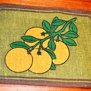 May include: A rectangular, green placemat with a brown border. The placemat features a branch of oranges with green leaves. The oranges are yellow with black outlines. The placemat is on a wooden surface.