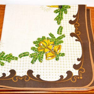 May include: A white tablecloth with a brown border and a Christmas design. The design features green pine branches, gold bells, and gold bows.