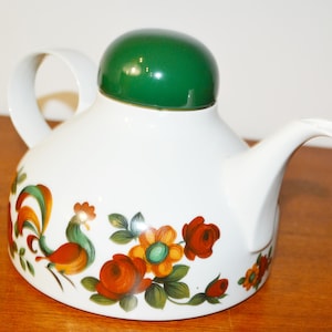 Puede incluir: Tetera de cerámica blanca con tapa verde y asa curva. La tetera está decorada con un gallo y un diseño floral en naranja, rojo, verde y amarillo. La tetera está sobre una superficie de madera.