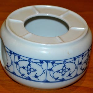 Puede incluir: Calienta-tetera de cerámica blanca con un diseño floral azul y blanco. El calienta-tetera tiene una abertura circular en el centro para una tetera.