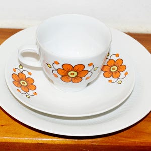 May include: A white teacup, saucer, and plate set. The cup and saucer feature orange floral designs. The set is on a wooden surface, creating a vintage aesthetic. The cup has a handle.