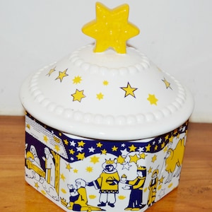 May include: A white ceramic jar with a lid, adorned with yellow stars and a yellow star-shaped handle. The jar's sides feature a blue and yellow illustration of the Three Wise Men and camels. The jar is approximately 5 inches tall.