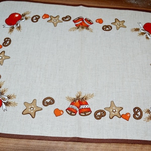 Puede incluir: Un mantel individual de lino beige con borde marrón presenta un diseño festivo de manzanas rojas, estrellas de jengibre, pretzels y corazones.