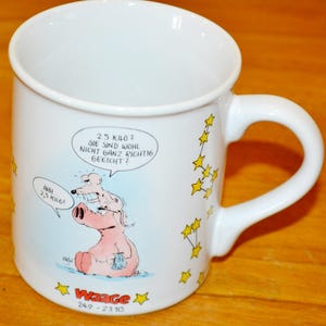 Puede incluir: Taza de cerámica blanca con una ilustración de dibujos animados de un cerdo y un ratón. La taza tiene un diseño de estrellas amarillas y el texto "25 KILO 2 SIE SIND WOHL NICHT GANZ RICHTIG GEEICHT?" y "AHH 2,5 KILO!" La taza también tiene el texto "Waage 249-2310".