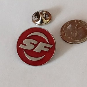 K&ouml;nnte beinhalten: Eine rote und silberne Anstecknadel mit den Buchstaben "SF" in einem wei&szlig;en Kreis.