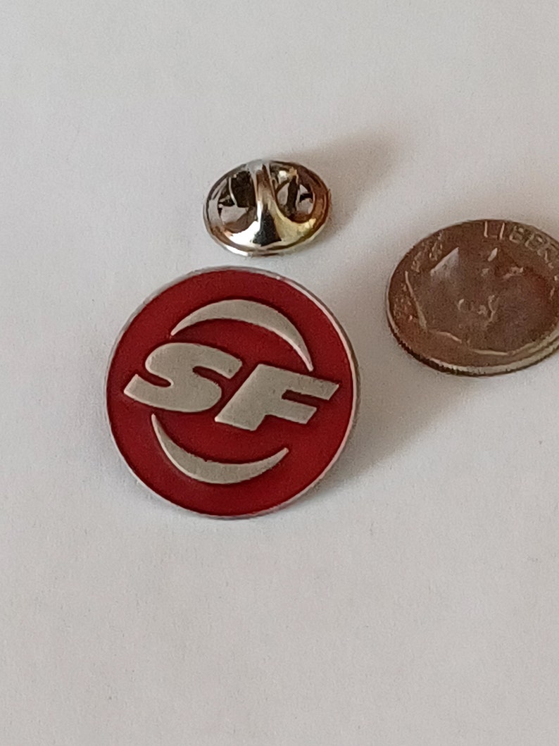 K&ouml;nnte beinhalten: Eine rote und silberne Anstecknadel mit den Buchstaben "SF" in einem wei&szlig;en Kreis.